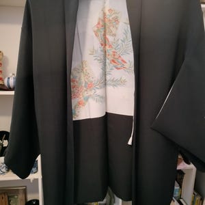May include: A dark gray kimono jacket with a white interior featuring a floral and fan design in shades of red, orange, and green. The jacket is hanging on a black hanger.