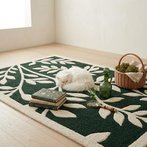 Op de afbeelding: Een rechthoekig vloerkleed met een donkergroene achtergrond en een groot, cr&egrave;mekleurig botanisch bladpatroon. Een witte kat ligt opgerold op het kleed, naast een stapel van twee boeken, een groene glazen karaf, een vergrootglas en een rieten mand.
