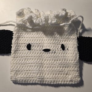 Op de afbeelding: Een witte gehaakte trekkoordtas met zwarte details. De tas heeft een schapen gezicht met zwarte ogen en een mond. Zwarte, ovale oren zijn aan de zijkanten bevestigd. De bovenkant van de tas is voorzien van een trekkoord.
