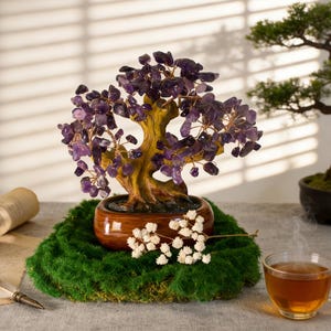 May include: A decorative amethyst crystal bonsai tree with a brown trunk and branches, set in a brown ceramic pot. The tree sits on a bed of green moss, with white flowers and a cup of tea in the background.