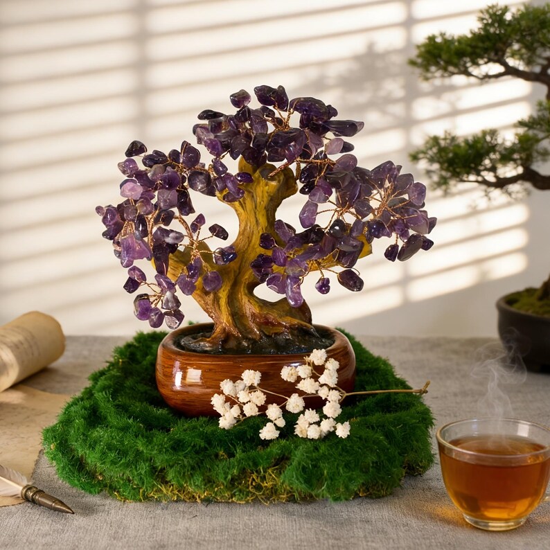 May include: A decorative amethyst crystal bonsai tree with a brown trunk and branches, set in a brown ceramic pot. The tree sits on a bed of green moss, with white flowers and a cup of tea in the background.