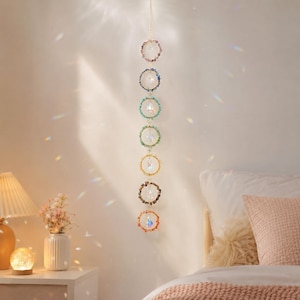 May include: A vertical hanging ornament featuring seven circular metal rings, each wrapped in small colorful beads. Each ring contains a clear, faceted crystal prism suspended in the center. The colors of the beads include purple, blue, teal, green, yellow, brown, and orange, arranged in a linear sequence.