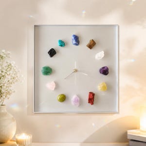Puede incluir: Un reloj de pared cuadrado con esfera gris claro y un marco plateado fino. En lugar de números tradicionales, doce piedras preciosas en bruto y multicolores están dispuestas en círculo para marcar las horas. Tres manecillas finas están centradas.