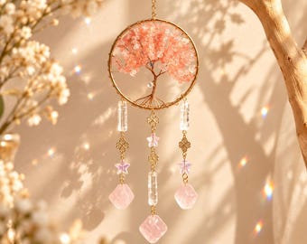 May include: A hanging ornament featuring a gold-toned circular frame containing a wire-wrapped tree with pink crystal leaves. Three dangling strands hang from the bottom, adorned with clear rectangular beads, gold clover spacers, star-shaped beads, and pink faceted crystal pendants.