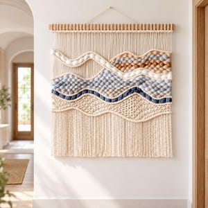 Puede incluir: Un gran tapiz de macramé rectangular colgado de un listón de madera. La pieza presenta patrones ondulados en capas de color crema, azul claro, azul oscuro y bronceado, creados con cordón anudado y texturas de fibra suave con flecos.