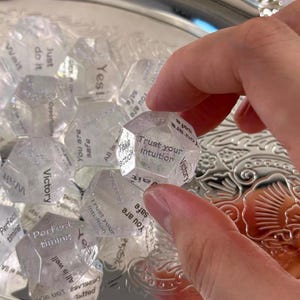 May include: A collection of clear, multi-faceted polyhedral dice featuring black printed text on each face. Visible phrases include Trust your intuition, Take action, You are safe, Victory, Wait, Perfect timing, Yes, and All is well.