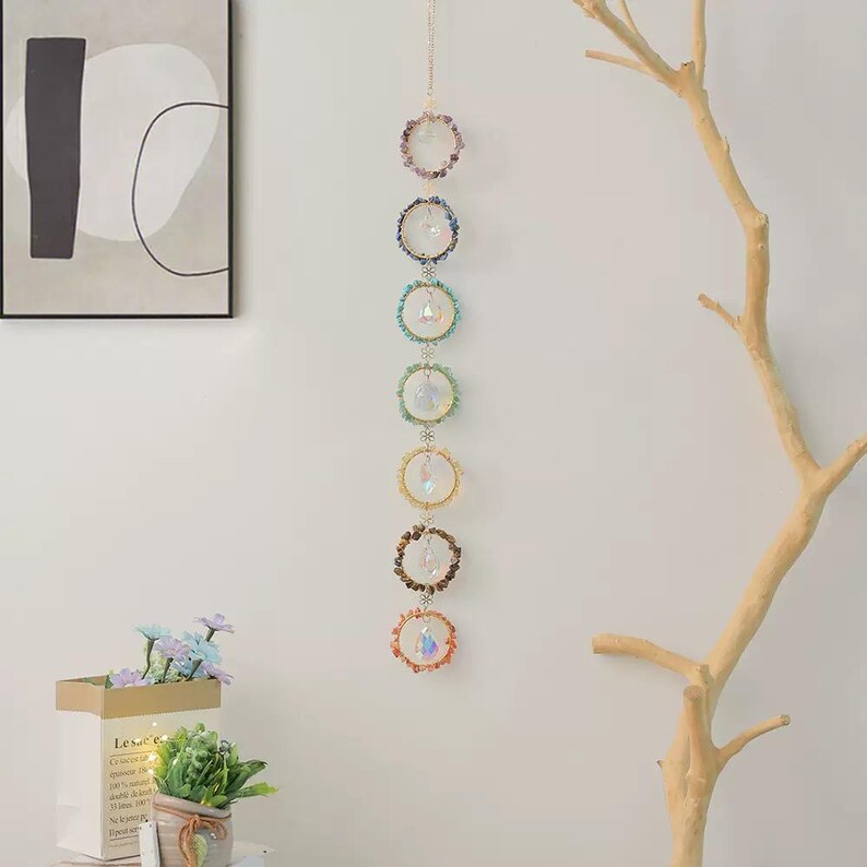 May include: A vertical hanging ornament featuring seven circular rings arranged in a column. Each ring is decorated with small colorful stones and holds a clear, faceted crystal prism in the center. The rings are connected by small metal links.