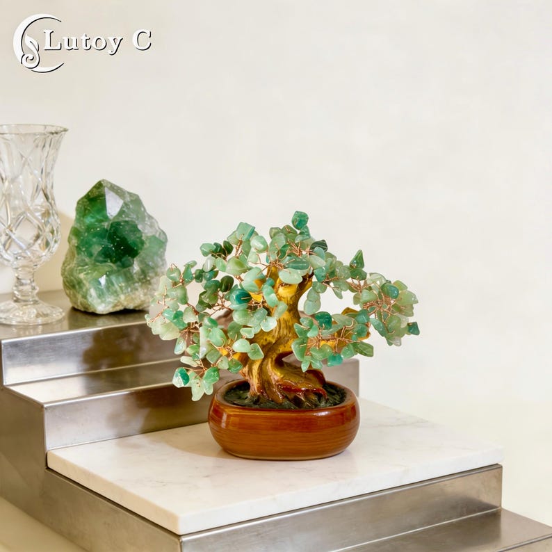 May include: A decorative bonsai tree with green gemstone leaves and a gold-colored trunk sits in a brown ceramic pot. The tree is on a white marble surface. A clear glass vase and a green crystal are in the background.