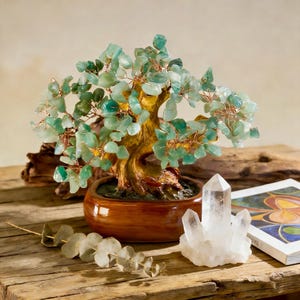 May include: A decorative bonsai tree with green gemstone leaves and copper wire branches sits in a brown ceramic pot. A clear quartz crystal and a book with colorful illustrations are next to the tree on a wooden surface.