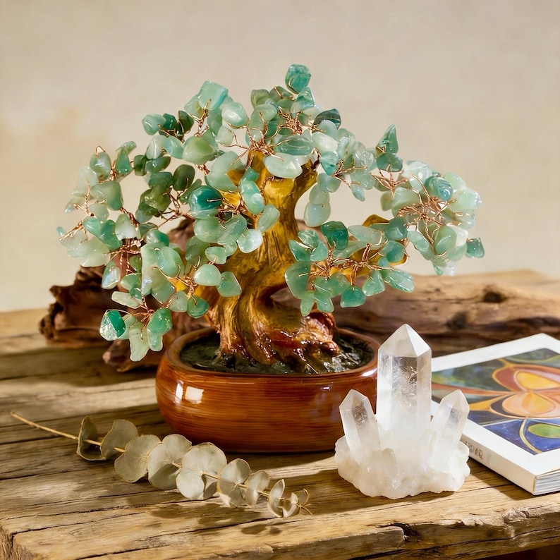 May include: A decorative bonsai tree with green gemstone leaves and copper wire branches sits in a brown ceramic pot. A clear quartz crystal and a book with colorful illustrations are next to the tree on a wooden surface.