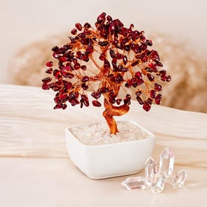 May include: A decorative tree sculpture with a copper wire trunk and branches, adorned with deep red gemstones. The tree sits in a square white ceramic pot filled with white stones. Clear quartz crystals are arranged nearby.
