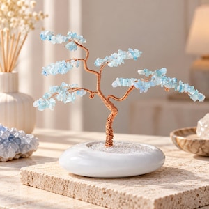 May include: A decorative tree sculpture featuring a twisted copper wire trunk and branches adorned with small, light blue tumbled gemstone leaves. The tree is anchored in a smooth, white, oval-shaped ceramic base filled with clear crystals.