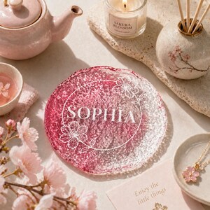 Könnte beinhalten: Ein rosa-weißer Glasuntersetzer mit dem Namen "SOPHIA" in weißer Schrift, umgeben von floralen Designs. Der Untersetzer ist Teil einer Kollektion, die eine Teekanne, eine Teetasse, eine Kerze und einen Diffusor umfasst, alle mit floralem Thema.
