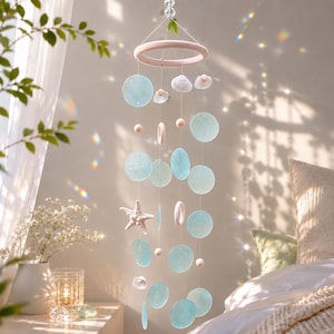 May include: A hanging mobile featuring a pale pink circular frame suspended by clear beads. It includes light blue translucent glass discs, white seashell shapes, a white starfish, and small wooden beads hanging on thin strings from the frame.