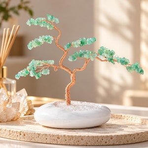 Puede incluir: Una escultura decorativa de árbol hecha de alambre con un tronco y ramas de metal color cobre trenzado, adornada con pequeñas hojas de piedras preciosas pulidas de color verde claro sobre una base de piedra blanca ovalada.