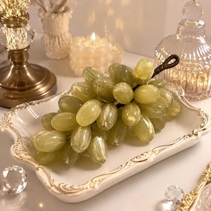 Puede incluir: Un racimo de uvas artificiales de color verde claro translúcido con un tallo marrón oscuro, sobre una bandeja rectangular de cerámica blanca con un borde festoneado adornado con detalles dorados. Las uvas tienen un acabado liso y brillante.