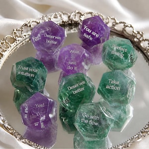 May include: A collection of multi-faceted purple and green gemstone dice featuring engraved white text including phrases such as You are safe, Trust your intuition, Wait just do it, Deserves better, Perfect timing, Take action, and Go with the flow.