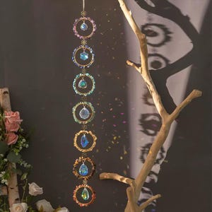 May include: A vertical hanging ornament featuring seven gold-toned metal rings arranged in a column. Each ring is decorated with small, multi-colored raw stones and contains a central clear, faceted crystal prism that reflects light.