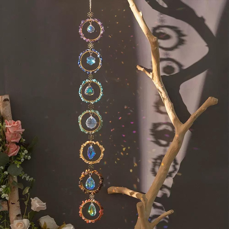 May include: A vertical hanging ornament featuring seven gold-toned metal rings arranged in a column. Each ring is decorated with small, multi-colored raw stones and contains a central clear, faceted crystal prism that reflects light.