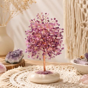 Könnte beinhalten: Eine dekorative Baumskulptur mit zahlreichen kleinen, polierten violetten Amethyst-Edelsteinen, die an dünnen, kupferfarbenen Drahtzweigen befestigt sind. Der Baum hat einen kupferfarbenen Stamm und steht auf einem runden, cremeweißen Steinsockel.