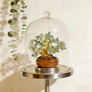 May include: A decorative bonsai tree sculpture with green gemstone leaves and a gold-colored trunk, housed under a clear glass cloche. The tree sits in a wooden pot on a silver metal stand, creating a unique home decor accent.