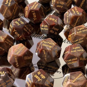 May include: A collection of brown, multi-faceted polyhedral dice featuring gold engraved text. Each face displays various phrases including Yes!, Just do it, Go with the flow, Trust your intuition, You are safe, All is well, Perfect timing, Wait, and Victory.