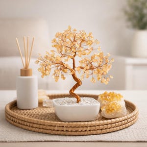 Könnte beinhalten: Ein dekorativer Drahtbaum mit bernsteinfarbenen Kristallen in einem weißen quadratischen Topf, der auf einem gewebten Tablett steht. Eine weiße Diffusorflasche mit Holzstäbchen und zwei rohe Citrin-Kristallcluster befinden sich ebenfalls auf dem Tablett.