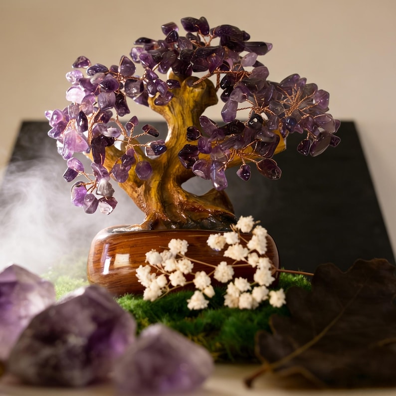 May include: A decorative amethyst gemstone tree with a wooden trunk and base. The tree's branches are made of copper wire and adorned with purple amethyst crystals. The tree sits on a bed of green moss, with white flowers and amethyst crystals in the foreground.