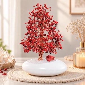 May include: A decorative tree sculpture featuring numerous small, polished red gemstone chips attached to twisted copper wire branches. The tree is anchored in a smooth, white, oval-shaped ceramic base filled with tiny clear crystals.