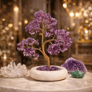 May include: A decorative tree sculpture featuring numerous small purple amethyst gemstone chips attached to twisted gold-toned wire branches. The tree is anchored in a round, smooth white stone base filled with loose purple amethyst stones.
