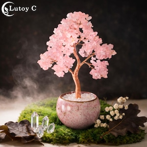 Könnte beinhalten: Eine dekorative Baumskulptur mit einem Kupferstamm und rosa Kristallblüten. Der Baum steht in einem rosa Keramiktopf, der mit weißen Steinen gefüllt ist und auf einem Bett aus grünem Moos steht. Klare Kristalle und Trockenblumen sind ebenfalls vorhanden.