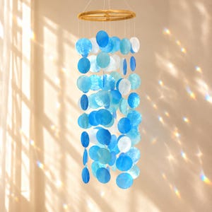 May include: A hanging mobile featuring a circular wooden frame with multiple strands of translucent, circular blue and white shell discs suspended by thin strings, creating a layered, cascading arrangement of varying shades of blue.