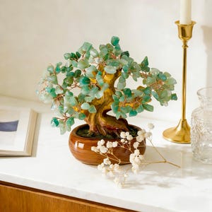 May include: A decorative bonsai tree with a brown ceramic pot. The tree features a textured trunk and branches adorned with green and blue-green gemstones. The tree is displayed on a white surface.