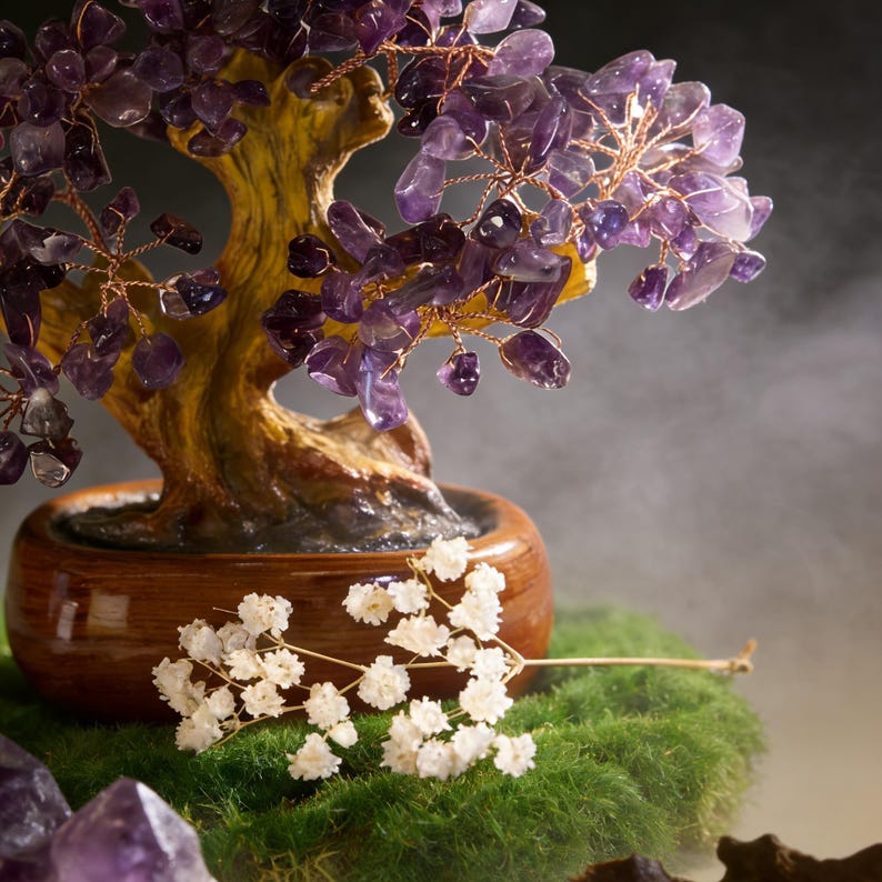 May include: A decorative amethyst crystal tree sculpture with a brown wooden trunk and branches. The tree is set in a small, round, brown pot and sits on a bed of green moss. White flowers are in the foreground.