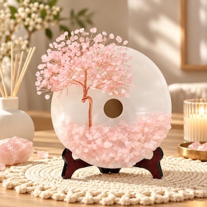 Könnte beinhalten: Eine dekorative Skulptur bestehend aus einer weißen, kreisförmigen Steinscheibe mit einem Loch in der Mitte, verziert mit einem drahtgewickelten Baum aus kleinen rosa Rosenquarzsplittern. Die Basis der Scheibe ist mit losen rosa Kristallen bedeckt.