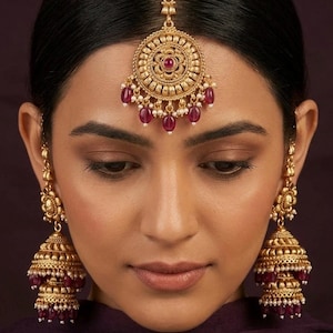 May include: A gold-toned maang tikka and matching jhumka earrings set. The circular maang tikka features a central floral motif with deep red stones and small white beads. The tiered bell-shaped earrings are adorned with red stones and white beads.