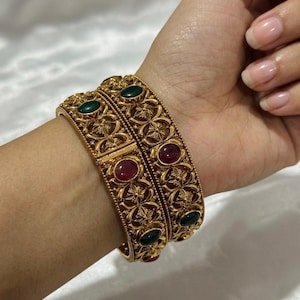 Puede incluir: Dos brazaletes de metal en tono dorado con un intrincado patrón de filigrana y piedras cabujón ovaladas incrustadas en colores rojo intenso y verde oscuro alternados. Los brazaletes tienen un diseño articulado y acabado metálico pulido.