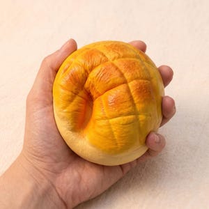 Op de afbeelding: Een ronde, geel en oranje broodje met een rasterpatroon bovenop, vastgehouden in een hand. Het brood heeft een zacht, getextureerd uiterlijk, met een kleurengradatie van lichtgeel tot diep oranje.