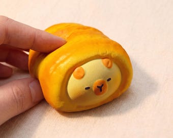 Pode incluir: Uma figura de urso amarela e fantasiosa com um exterior semelhante a um croissant. O urso tem um rosto amarelo claro com detalhes de orelhas laranja, olhos pretos e uma pequena boca marrom em forma de "Y". A figura é segurada por uma mão.