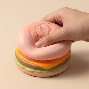 Op de afbeelding: Een roze, squishy burger speelgoed met een beer gezicht in reliëf op de bovenste broodje. De burger heeft lagen tomaat, kaas en sla. Een hand knijpt in het bovenste broodje, waardoor het speelgoed wordt samengedrukt.