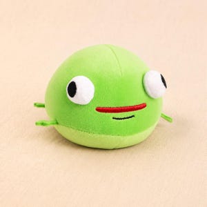 May include: A round, lime green plush toy shaped like a blob. It features two large, white, circular eyes with black pupils, a horizontal red embroidered mouth, and a thin black embroidered line below it. Small green limbs extend from the sides.