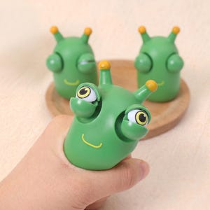 May include: Green alien-like toy with yellow accents, held in a hand. The toy has large, expressive eyes and a smiling mouth. Two additional toys are visible in the background, resting on a wooden plate.