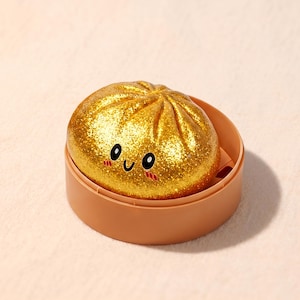Könnte beinhalten: Ein goldfarbener, glitzernder Knödel mit einem lächelnden Gesicht, der in einem braunen Dampfkorb liegt. Der Knödel hat eine strukturierte Oberfläche und schwarze Augen. Der Hintergrund ist hellpfirsichfarben.