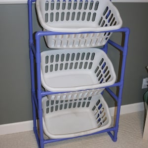May include: A three-tiered laundry sorter with a purple frame and three white plastic baskets. Each basket has a rectangular shape with numerous vertical slots for ventilation. The sorter is designed for organizing laundry.