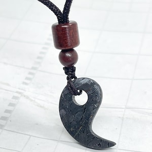 May include: A dark gray, textured, comma-shaped pendant with a hole near the top, suspended from a black cord. The cord features two smooth, cylindrical and spherical dark brown wooden beads above the pendant, creating a simple necklace.