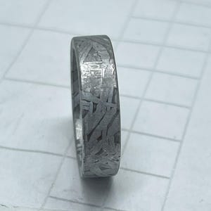 Peut inclure: Gros plan d'une bague argentée avec un motif texturé unique. La bague a un design à bande plate et un motif détaillé et entrelacé sur sa surface. La bague est probablement en métal et est présentée sur une surface blanche.
