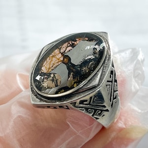 Peut inclure: Bague en argent avec une monture ovale en pierre transparente. La pierre contient un motif sombre et abstrait avec des touches d'or et de brun. La bague a un motif géométrique sur le bracelet et une face carrée.