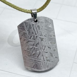 May include: A rectangular pendant with a unique, patterned surface, suspended from a light green cord. The pendant is a silver-gray color and has a textured appearance. The cord is attached to a silver-colored metal loop at the top of the pendant. The pendant is a dog tag style.