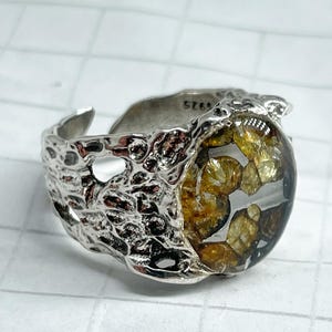 May include: A silver-toned ring with a textured band and a clear, oval-shaped center filled with amber-colored stones. The ring has a unique, organic design, and the band is stamped with "925".