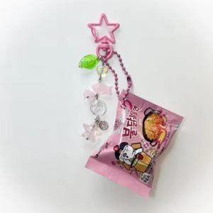 Pode incluir: Um chaveiro rosa com um topo em forma de estrela, uma corrente e pingentes decorativos. Os pingentes incluem uma folha verde, contas transparentes, estrelas rosa e uma espiral prateada. Um pacote rosa de macarrão instantâneo está anexado.